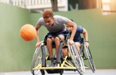 Two young men in sports wheelchairs chasing a basketball.