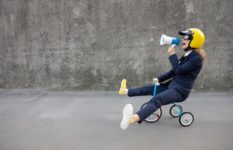 A woman riding a child's tricycle, wearing a helmet and shouting into a megaphone