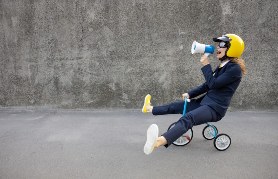 A woman riding a child's tricycle, wearing a helmet and shouting into a megaphone