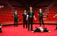 A scene from Julius Caesar: several men in suits surround a man on the floor who has been stabbed.