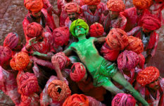A man covered in green colouring is carried on the shoulders of others wearing red turbans, seen from above. Jaipur, Rajasthan, India.