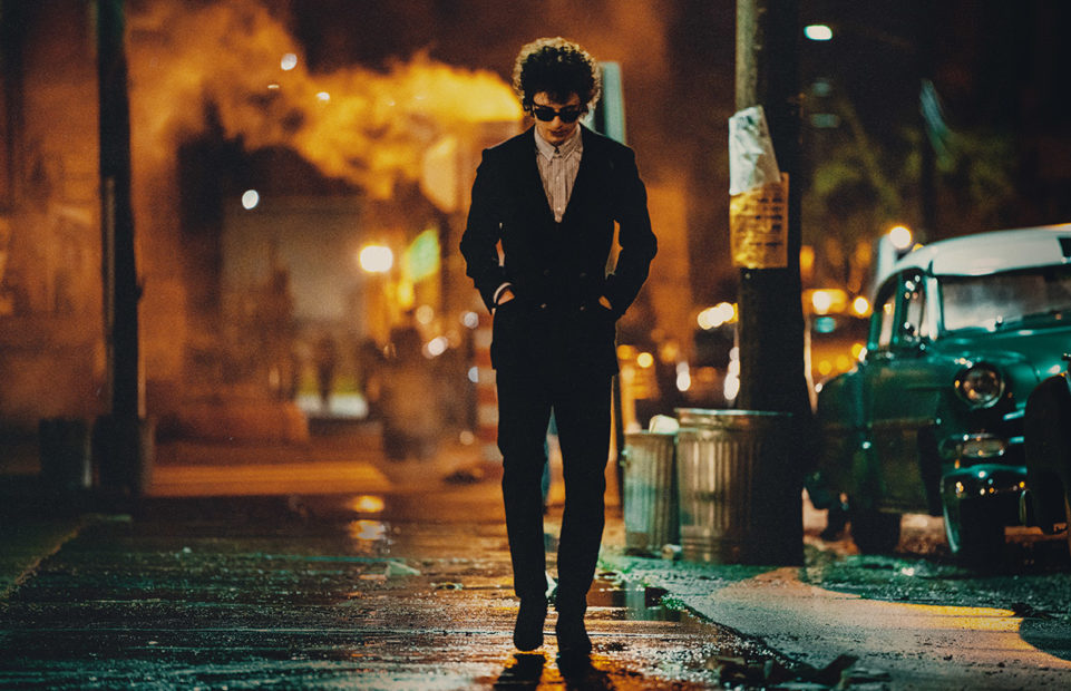 Timothée Chalamet as Bob Dylan, walking down a street dressed in black.