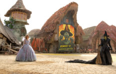 Glinda and Elphaba in a village with a large poster of the Wicked Witch of the West.