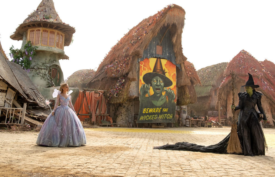 Glinda and Elphaba in a village with a large poster of the Wicked Witch of the West.